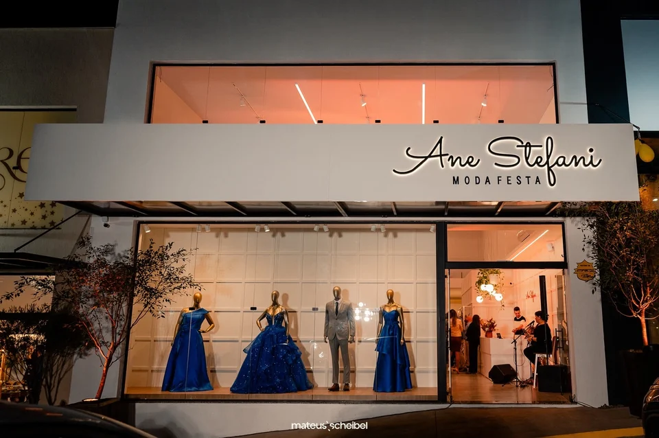 Fachada iluminada da Ane Stefani Moda Festa à noite, com vestidos e terno na vitrine